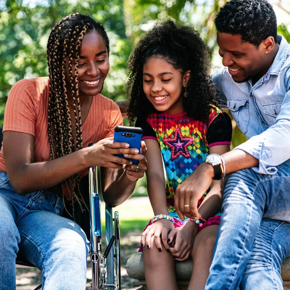 A woman in wheelchair holding a phone towards a young girl and gentleman whilst smiling